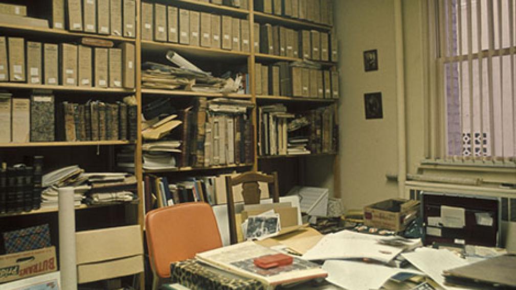 An archival image of an office with reference books and files on the desk