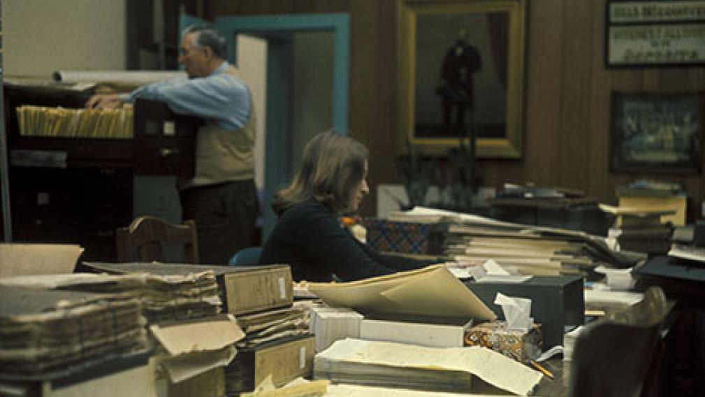 An archival image of two people in an office surrounded by files