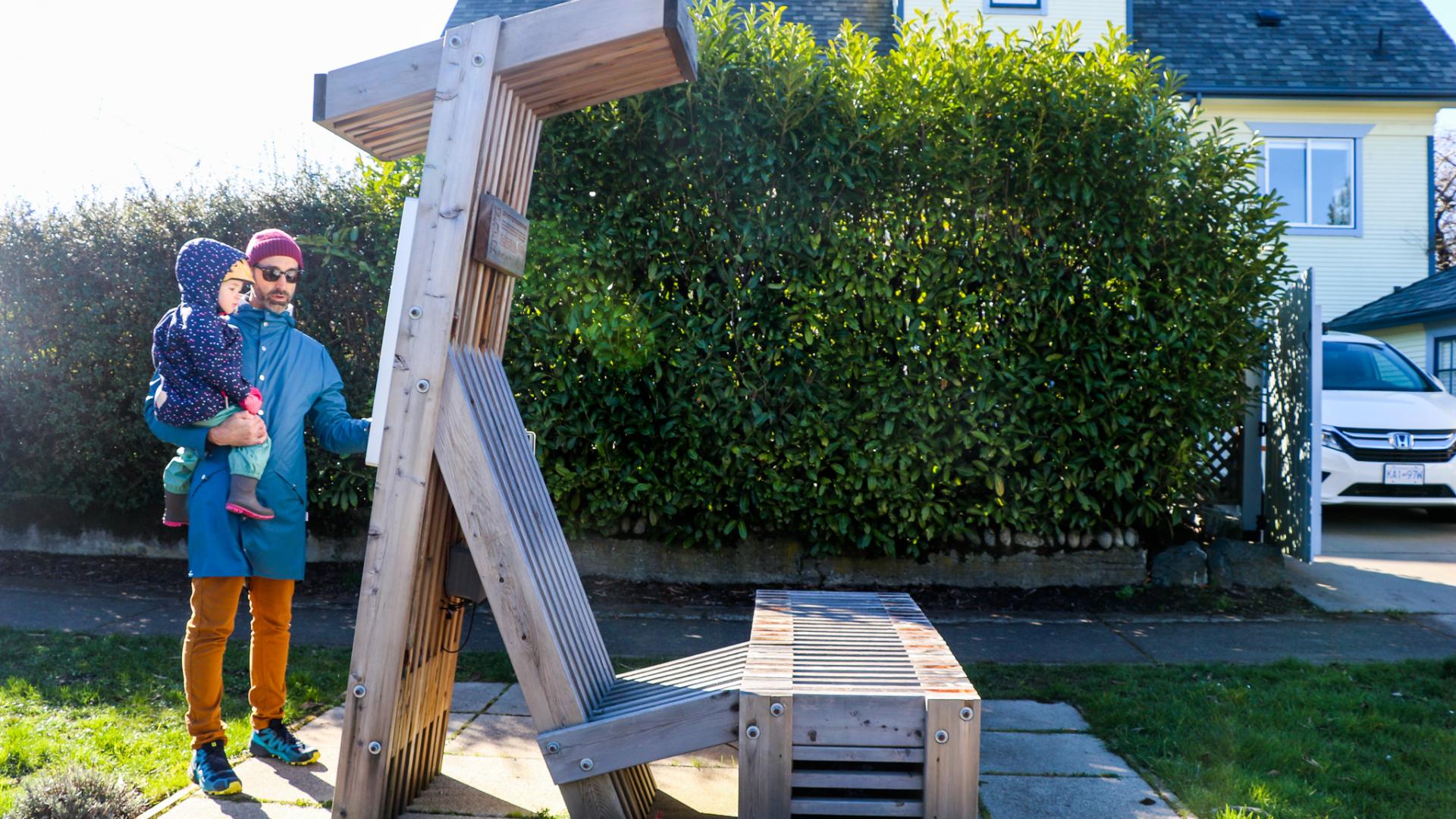 A father holds a child next to a large wood structure that resembles a large chair with a small canopy.