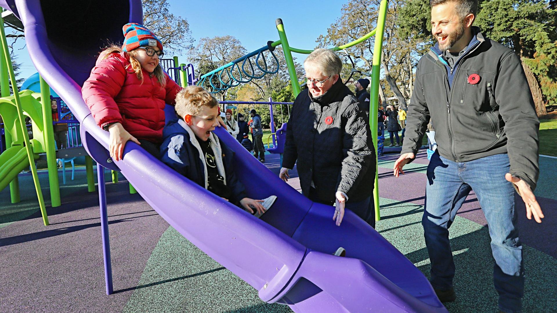 Photo of Mayor Alto and Derrick Newman, Acting Director of Parks, Recreation and Facilities, standing next to the new Stadacona Park playground while children enjoy sliding down the purple slide. 