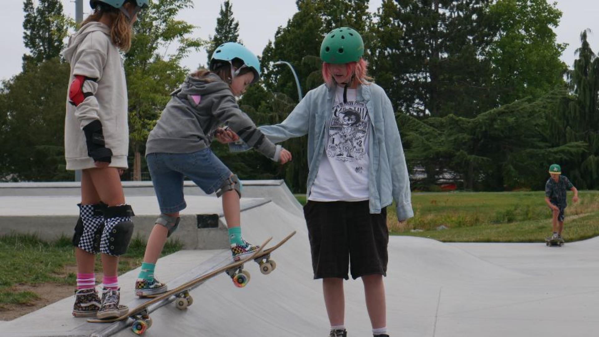 A skateboard instructor helps two students learn to drop into a skate bowl