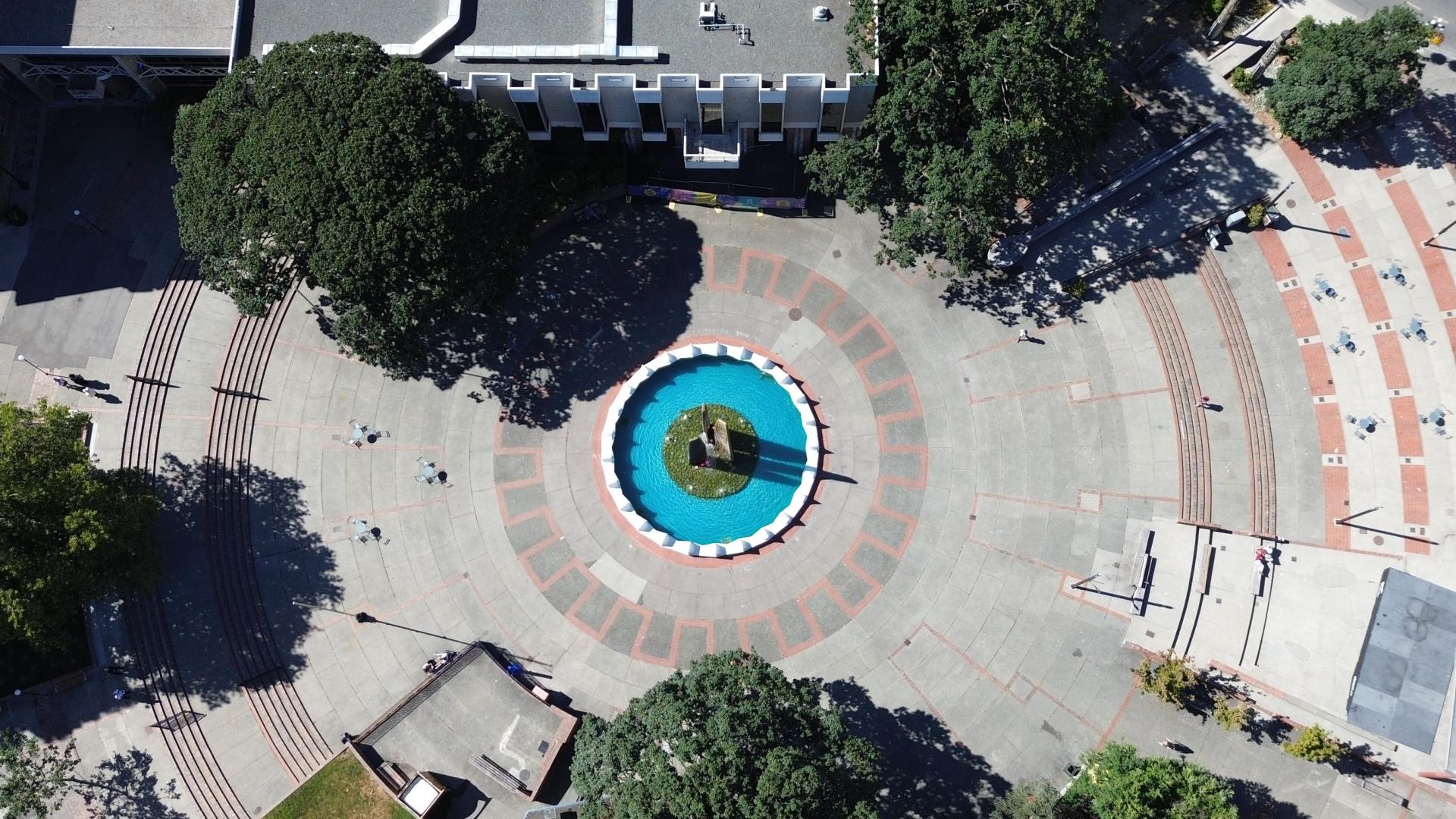 A bird's eye view of the current Centennial Square shows the central fountain water feature surrounded by concrete and trees. 