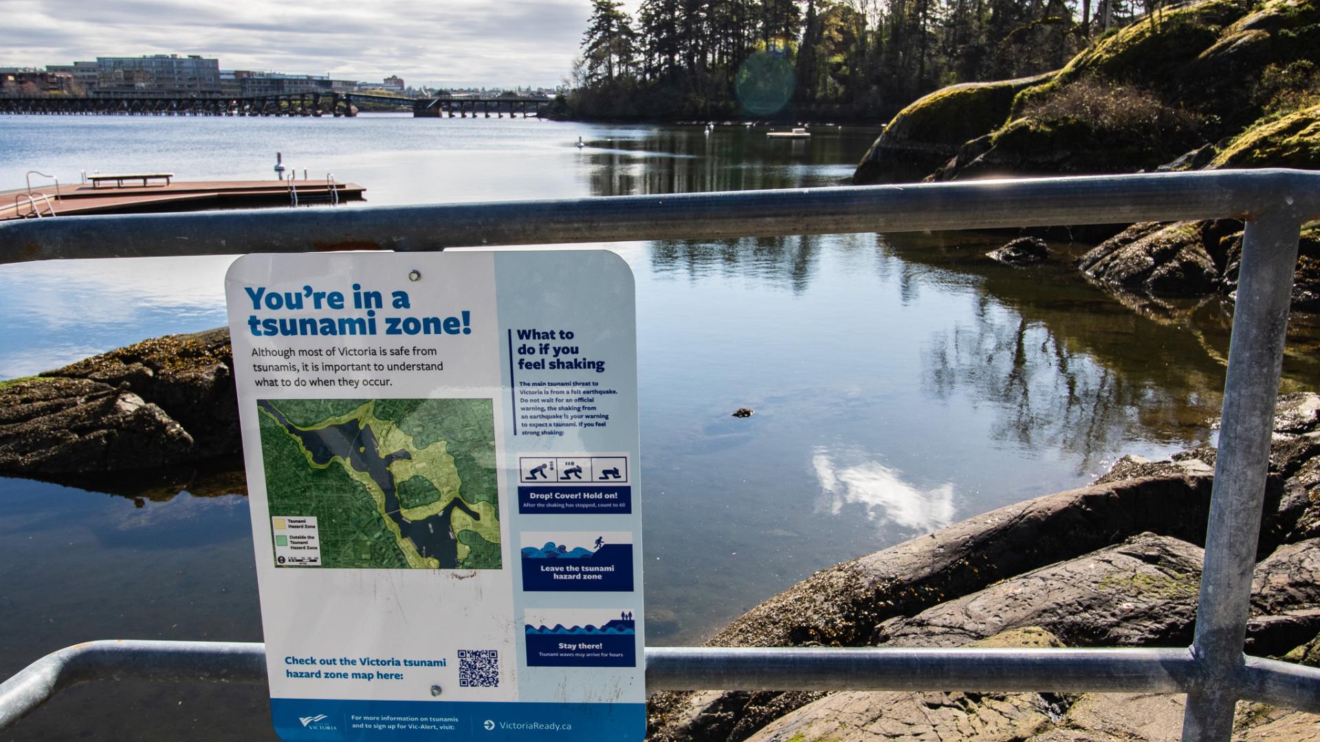 A tsunami hazard zone sign posted on the metal railing with Banfield dock and the Gorge Waterway in the background.