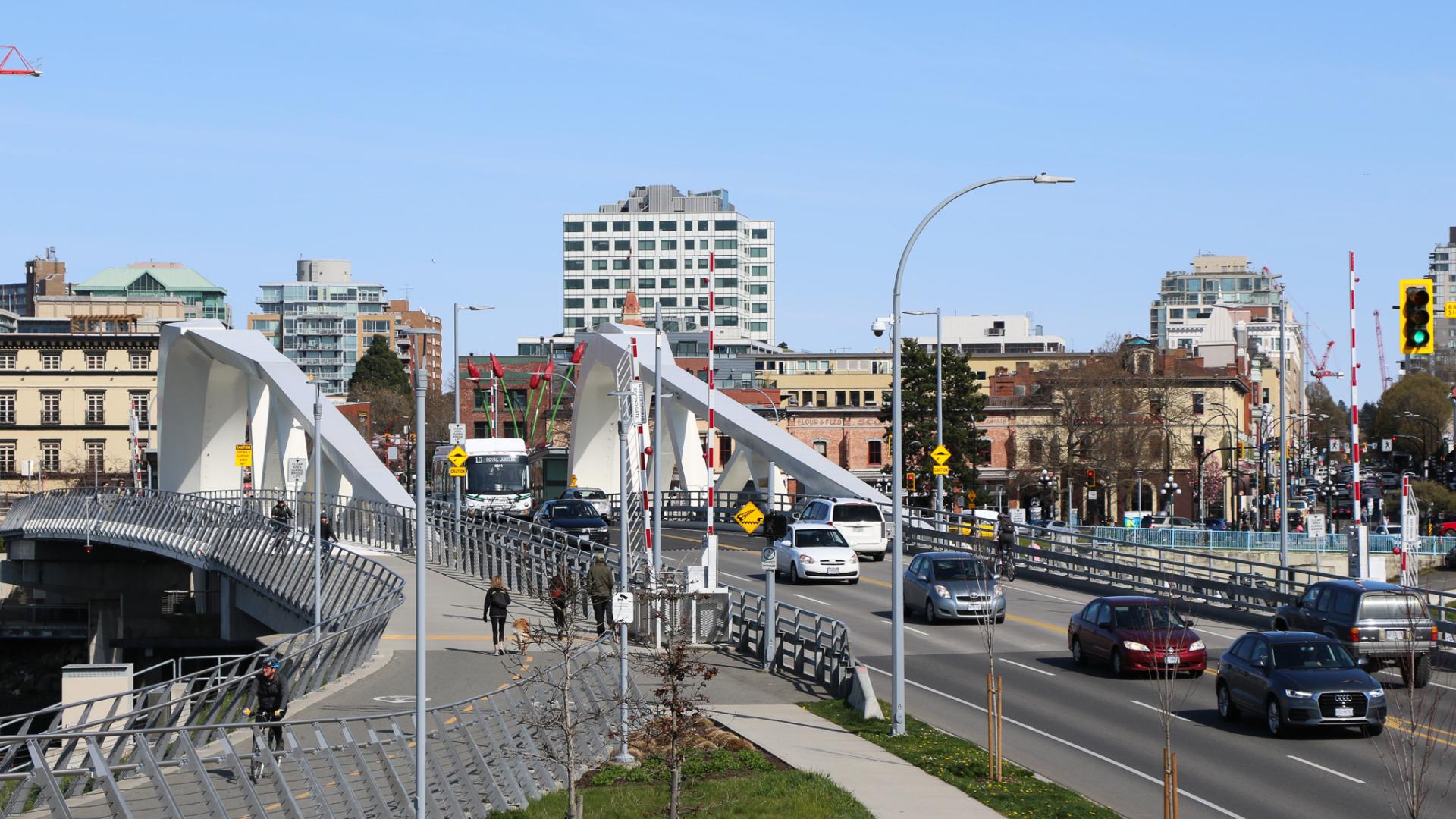 Johnson Street Bridge with pedestrians, cars and cyclists travelling over it, and downtown buildings visible in the background.
