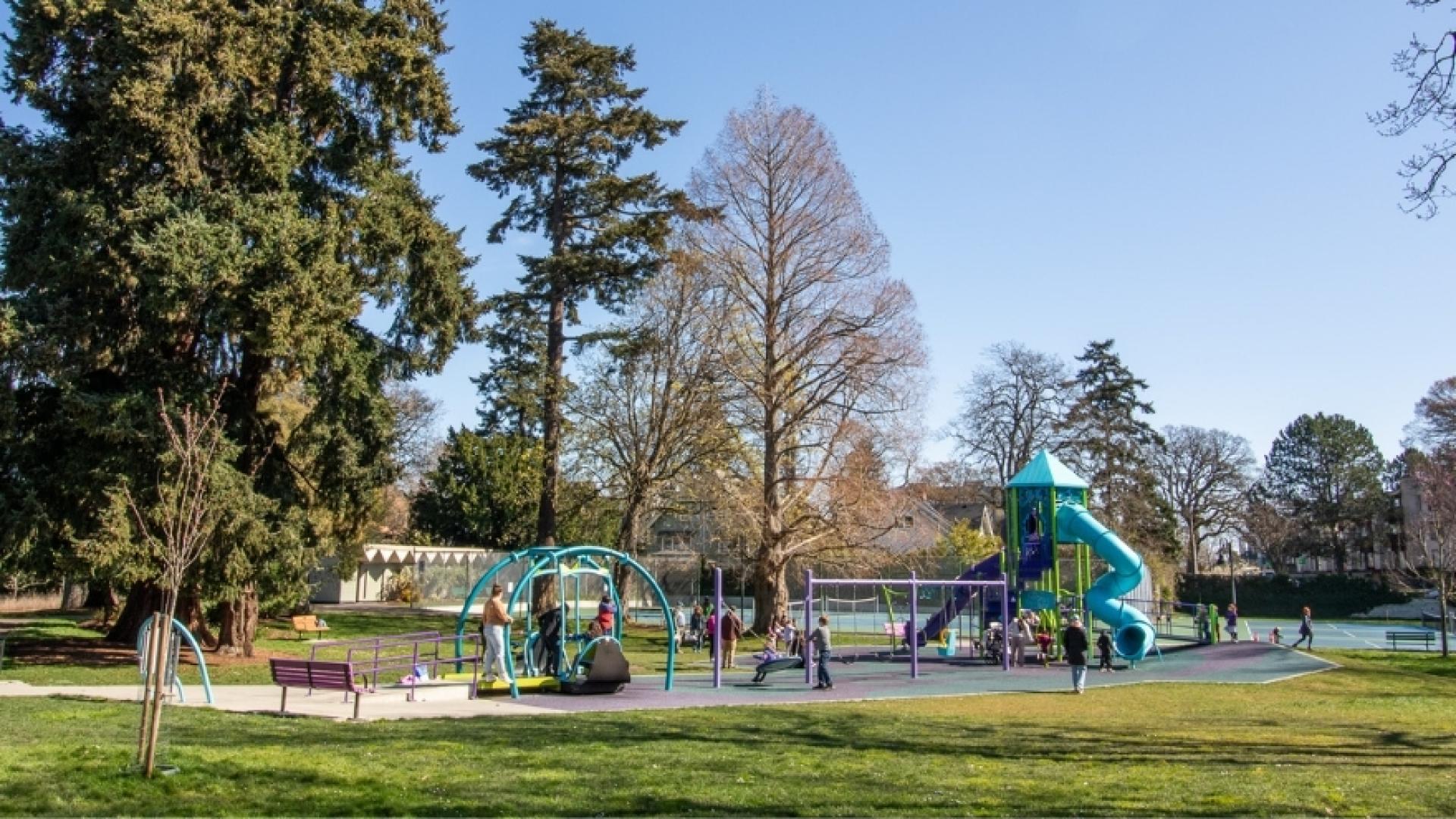 A playground in a park, featuring play structures, swings and climbing equipment. Several people are using the playground, which is surrounded by large trees and open grassy areas under a clear sky.