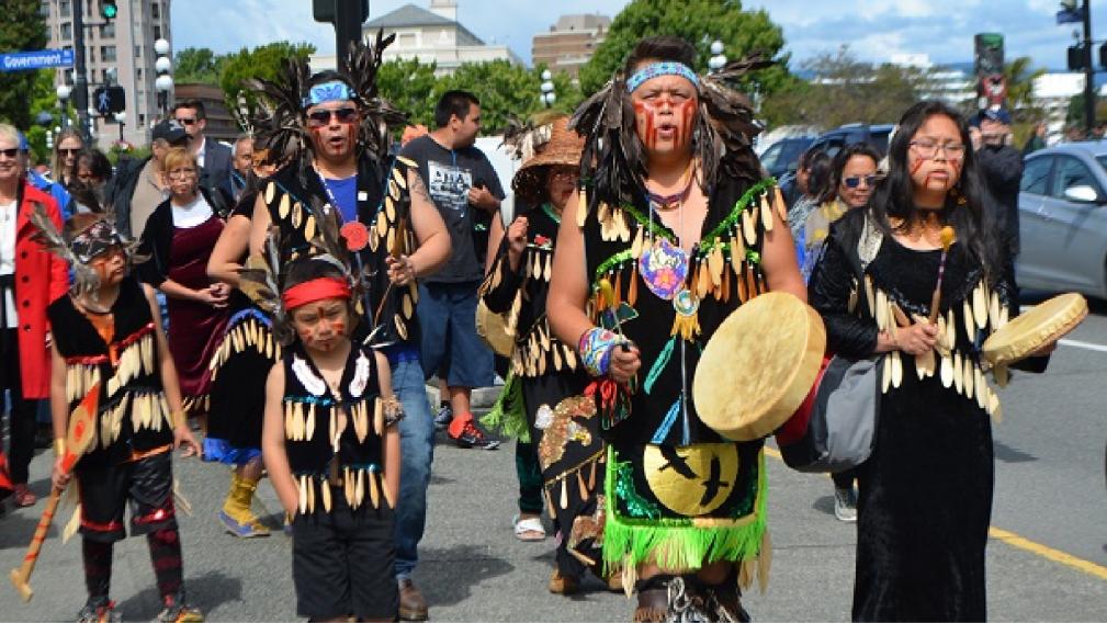 Image of members of the Songhees Nation and Esquimalt Nation in traditional ceremony clothing walking and drumming in a procession along Belleville Street