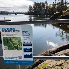 A tsunami hazard zone sign posted on the metal railing with Banfield dock and the Gorge Waterway in the background.