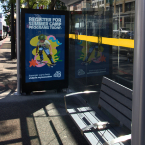 A new bus shelter on Douglas Street with glass walls, a bench and a digital screen advertising Summer Camp Registration.